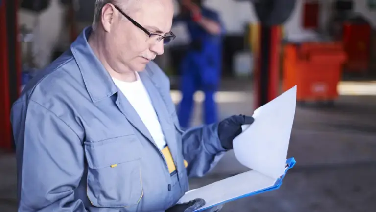 Een autotechnicus die u een garage werkt en een document op een klembord controleert.